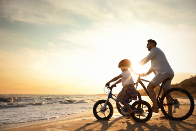 Happy father with son riding bicycles on sandy beach near sea at sunset