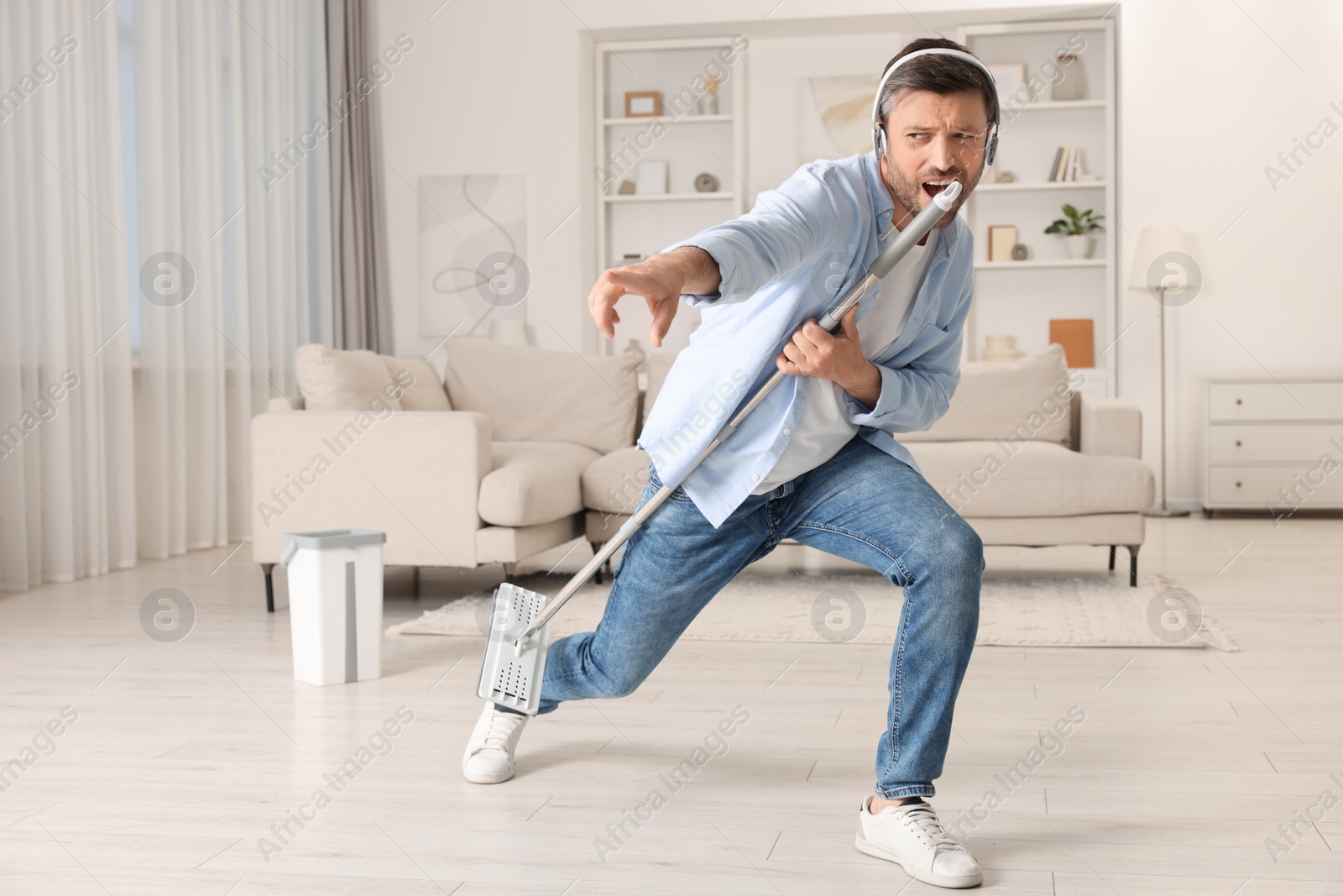 Photo of Enjoying cleaning. Happy man in headphones with mop singing while tidying up at home