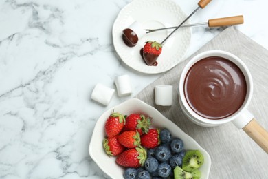 Fondue pot with melted chocolate, sweet marshmallows, fresh kiwi, different berries and forks on white marble table, flat lay. Space for text