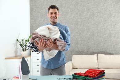 Photo of Man holding heap of clothes near ironing board at home. Space for text