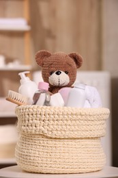 Knitted basket with baby cosmetic products, bath accessories and toy bear on white table indoors