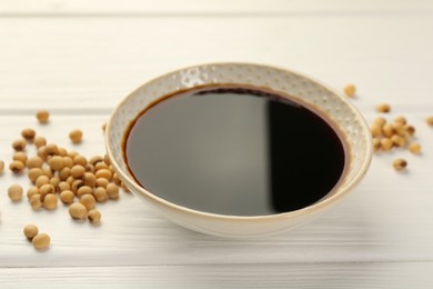 Photo of Tasty soy sauce in bowl and soybeans on white wooden table, closeup