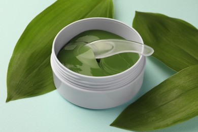 Photo of Jar of under eye patches with spoon and green leaves on turquoise background, closeup. Cosmetic product