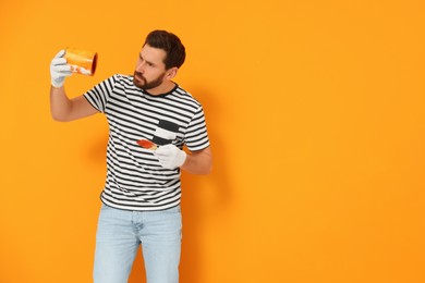 Photo of Confused designer with can of paint and brush near freshly painted orange wall, space for text