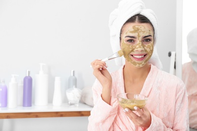 Young woman applying clay mask on her face in bathroom, space for text. Skin care