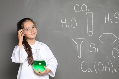 Photo of Little school child in laboratory uniform with flask of liquid and chemical formulas on grey background