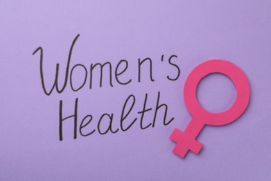 Photo of Female gender sign near text Women's Health on violet background, top view