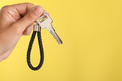 Photo of Woman holding key with black leather keychain on yellow background, closeup. Space for text
