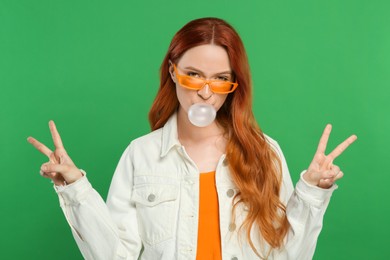 Photo of Beautiful woman in sunglasses blowing bubble gum and showing peace gesture on green background
