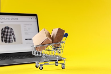 Photo of Online store. Mini shopping cart, parcels and laptop on yellow background, selective focus. Space for text