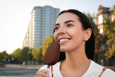 Photo of Beautiful young woman eating ice cream glazed in chocolate on city street