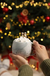 Photo of Woman holding cup of delicious cocoa with marshmallows near Christmas tree at home, closeup. Space for text