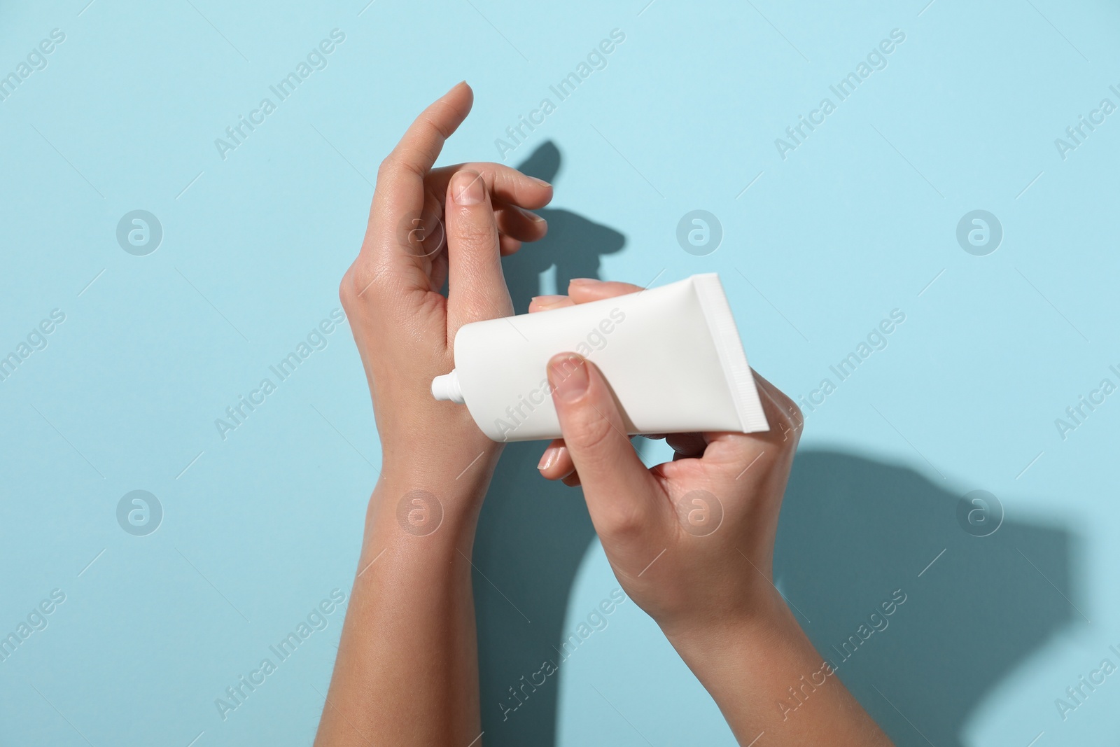 Photo of Woman applying cosmetic cream from tube onto her hand on light blue background, top view