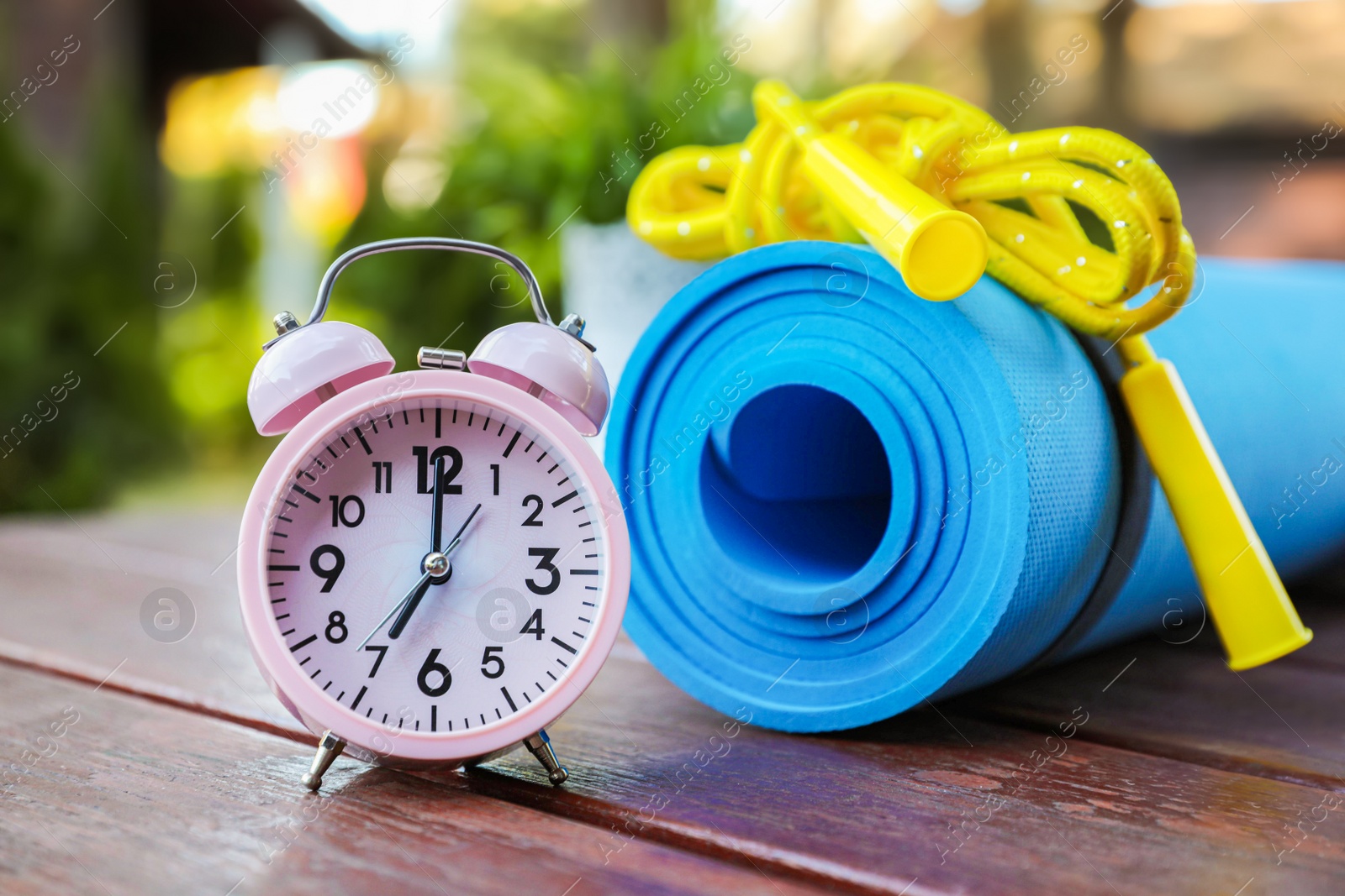 Photo of Alarm clock, fitness mat and skipping rope on wooden table outdoors. Morning exercise