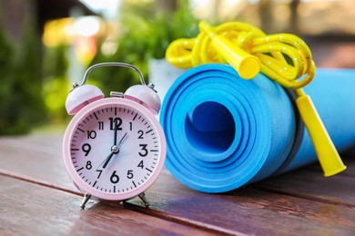Photo of Alarm clock, fitness mat and skipping rope on wooden table outdoors. Morning exercise