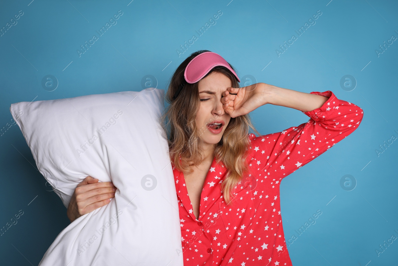 Photo of Young tired woman with sleeping mask and pillow yawning on light blue background