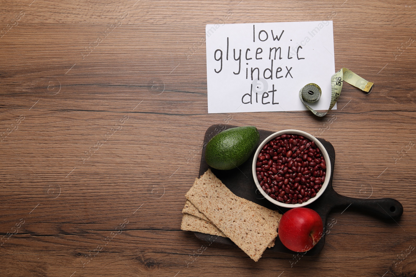 Photo of Paper with words Low Glycemic Index Diet, measuring tape and healthy products on wooden table, flat lay. Space for text