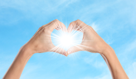 Image of Woman making heart around bright light with hands, closeup. Solar energy concept