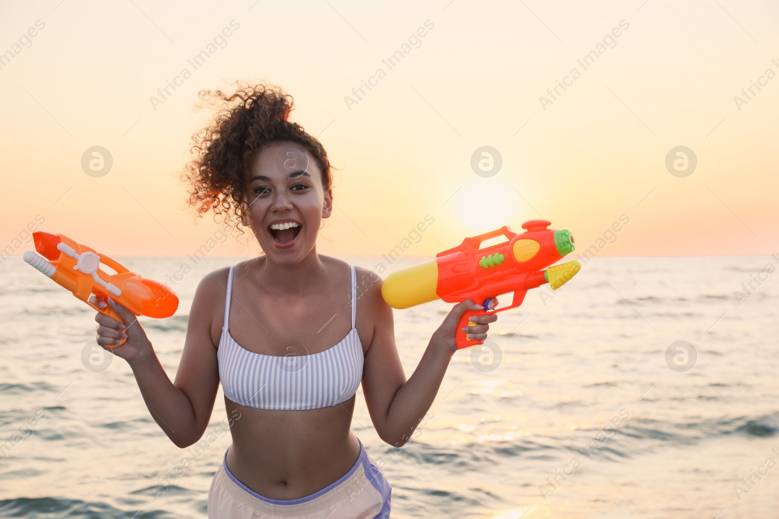 Photo of African American woman with water guns having fun on beach at sunset