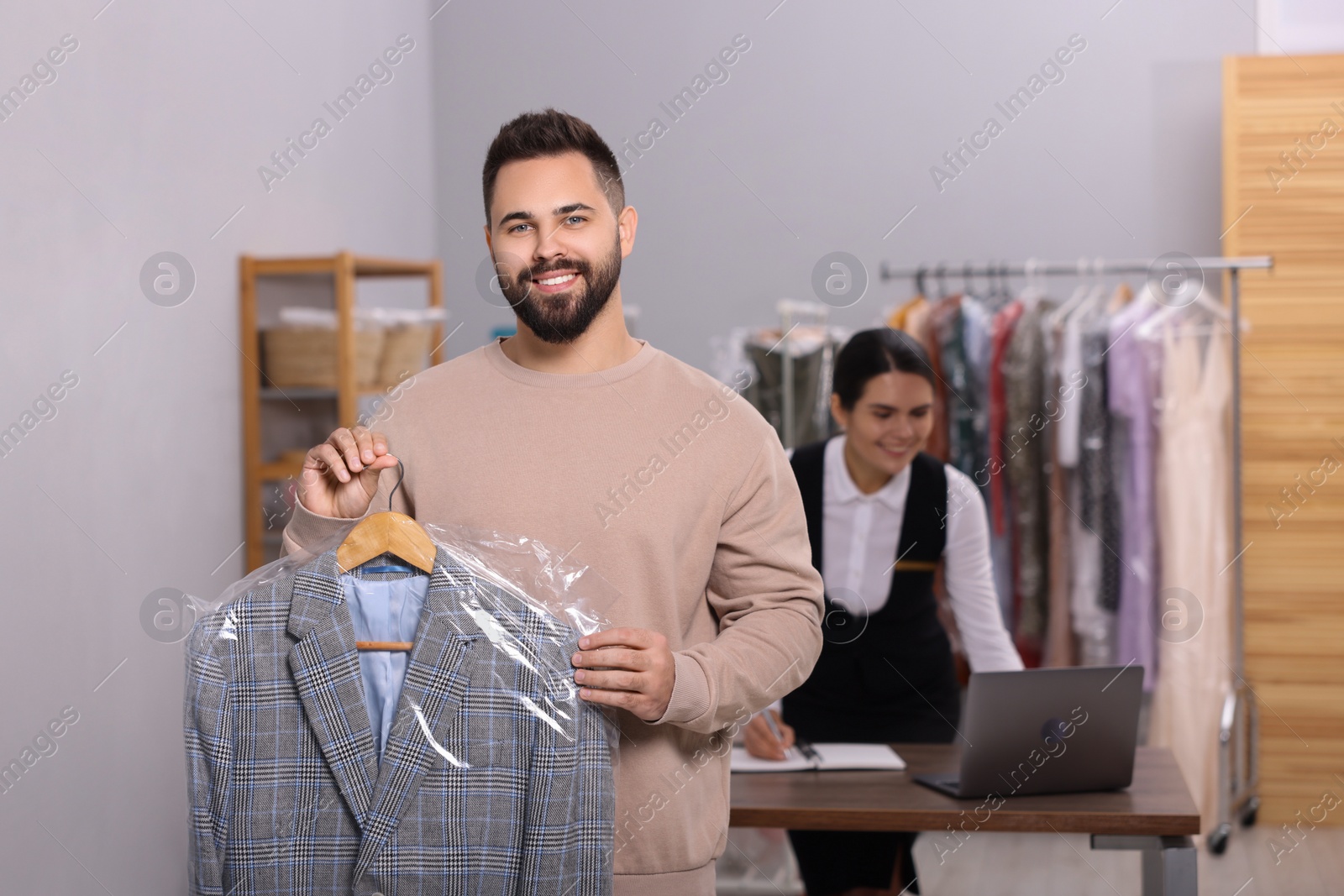 Photo of Dry-cleaning service. Happy man holding hanger with jacket indoors. Worker taking notes at workplace