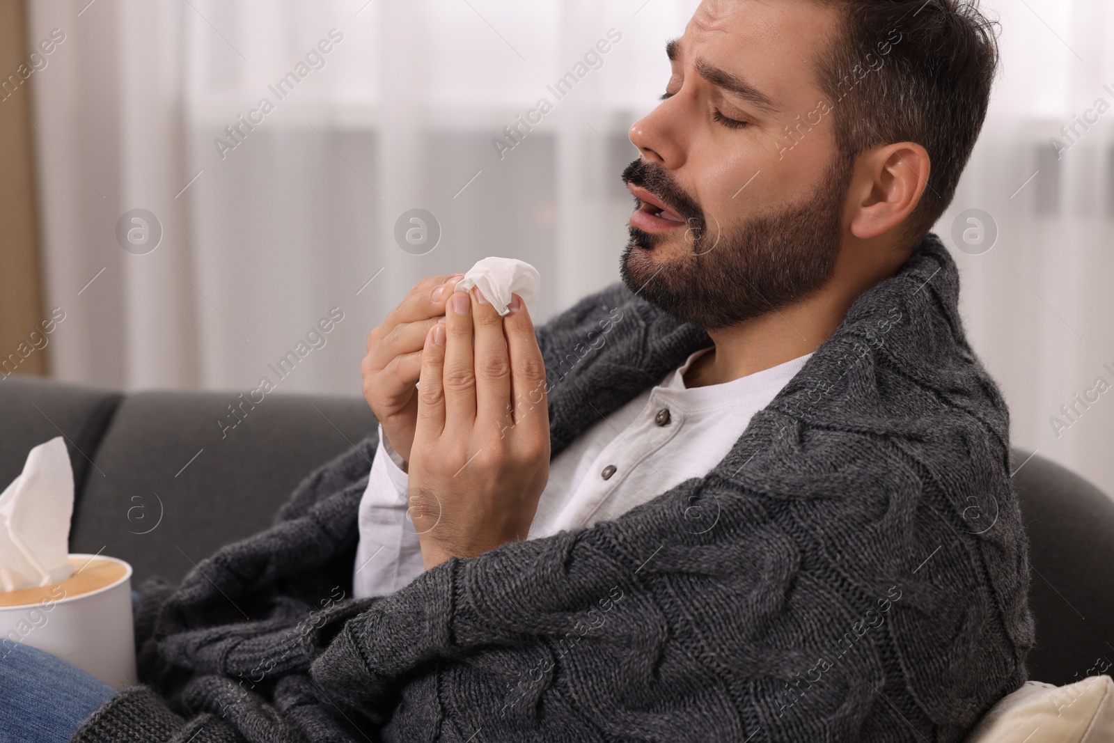 Photo of Sick man wrapped in blanket with tissue on sofa at home. Cold symptoms