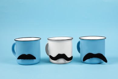 Cups with fake mustaches on light blue background