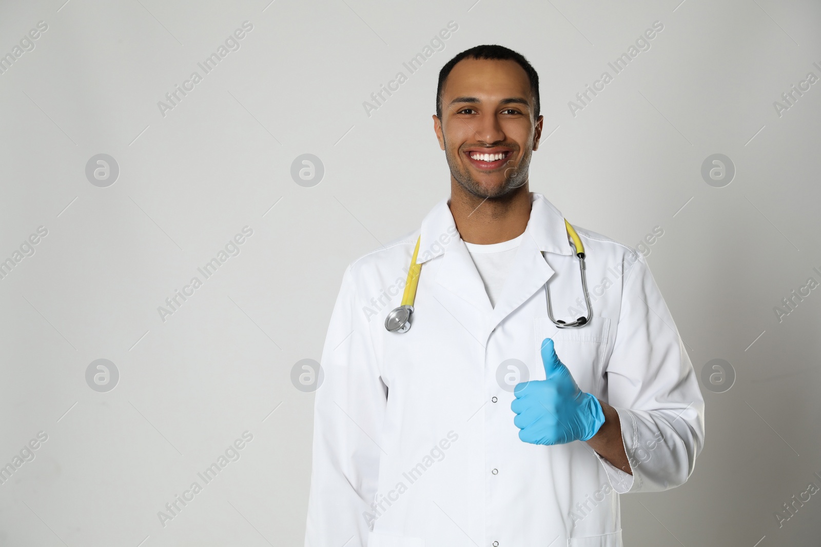 Photo of Doctor or medical assistant (male nurse) in uniform showing thumb up on light grey background. Space for text