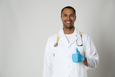 Photo of Doctor or medical assistant (male nurse) in uniform showing thumb up on light grey background. Space for text