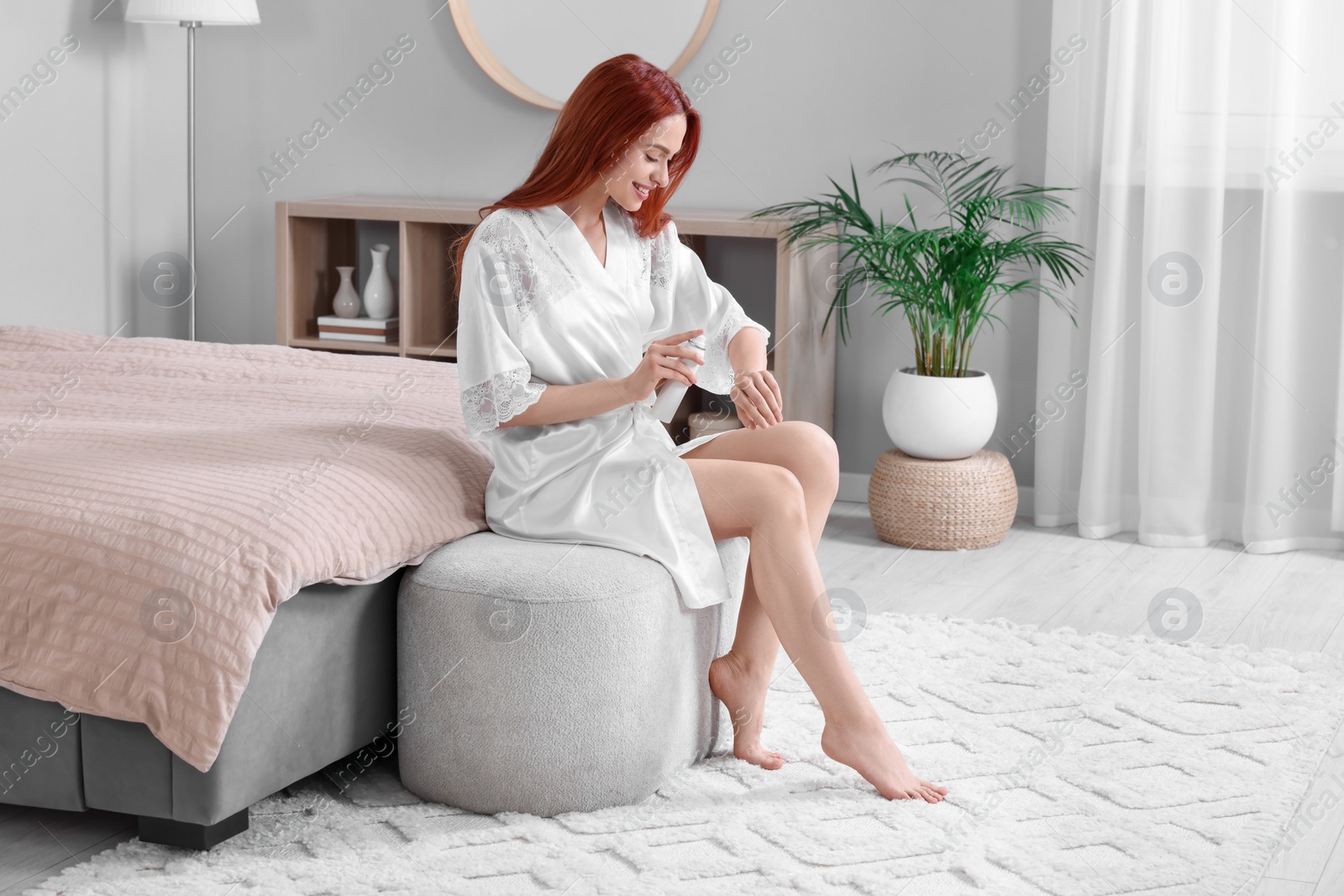 Photo of Beautiful young woman applying body spray onto arms in bedroom