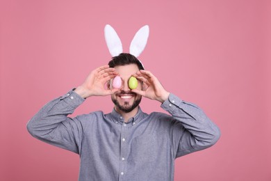 Happy man in bunny ears headband holding painted Easter eggs near his eyes on pink background