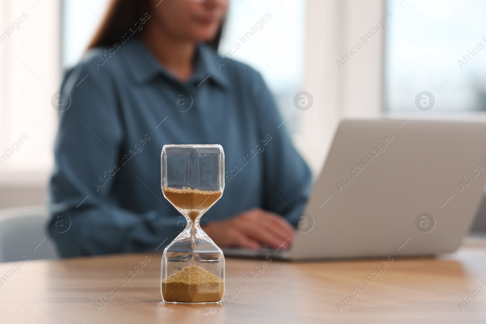 Photo of Hourglass with flowing sand on table. Woman using laptop indoors, selective focus