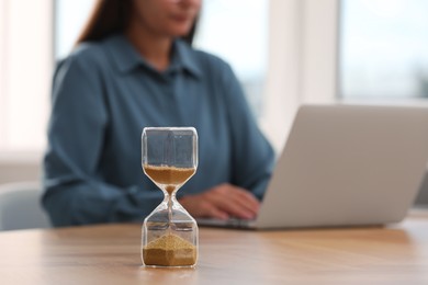 Hourglass with flowing sand on table. Woman using laptop indoors, selective focus