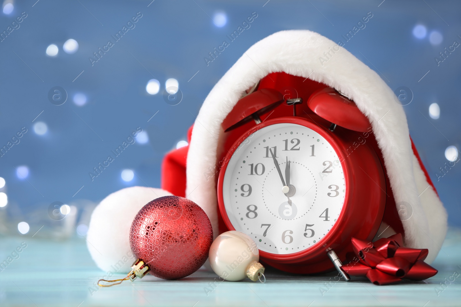 Photo of Alarm clock with decor on light blue wooden table against blurred Christmas lights, closeup. New Year countdown