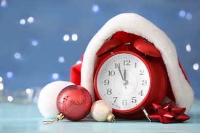 Photo of Alarm clock with decor on light blue wooden table against blurred Christmas lights, closeup. New Year countdown