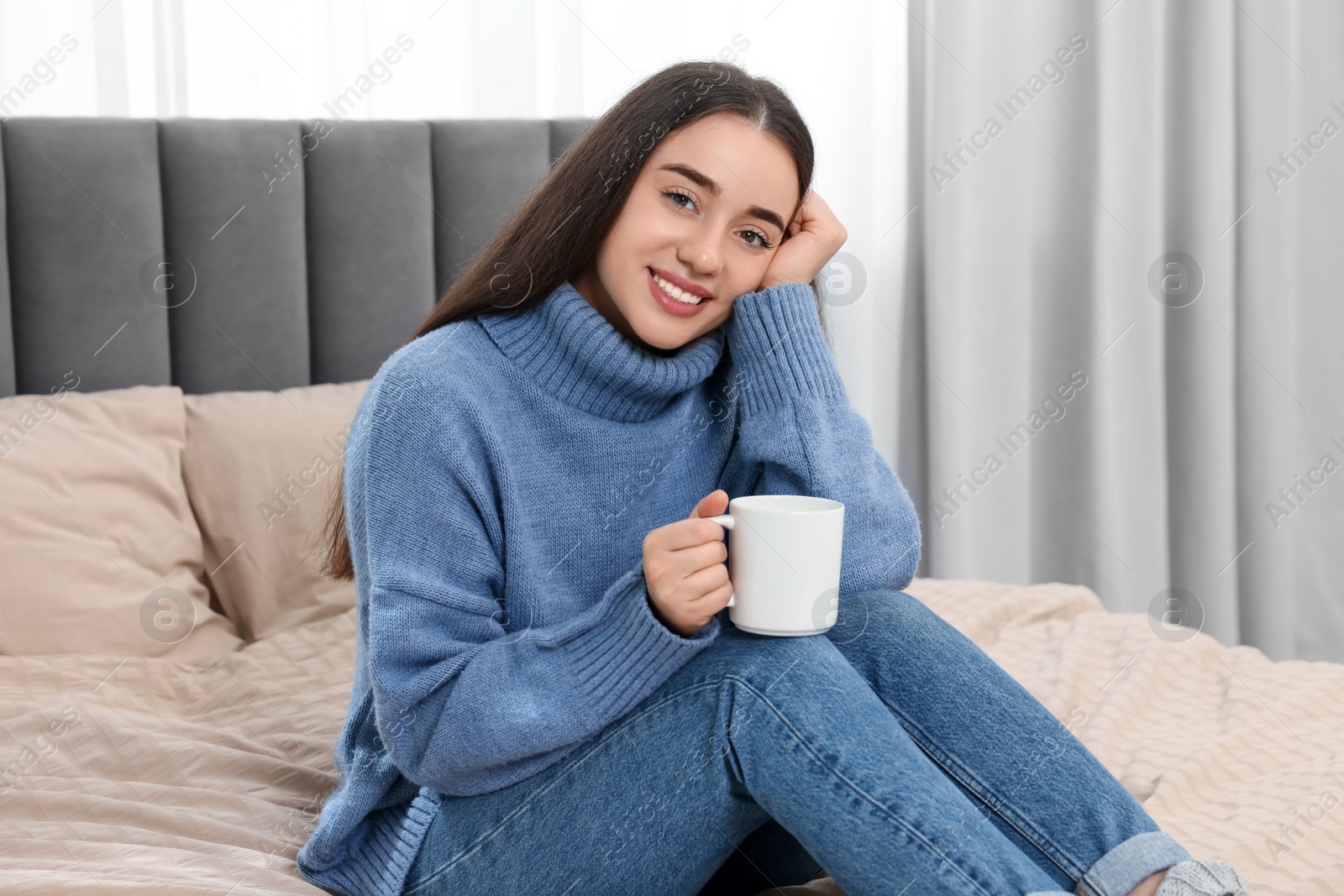 Photo of Happy young woman holding white ceramic mug on bed at home