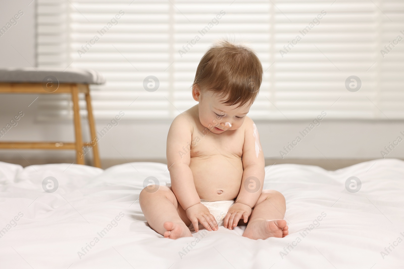 Photo of Cute little baby with moisturizing cream on body sitting on bed at home