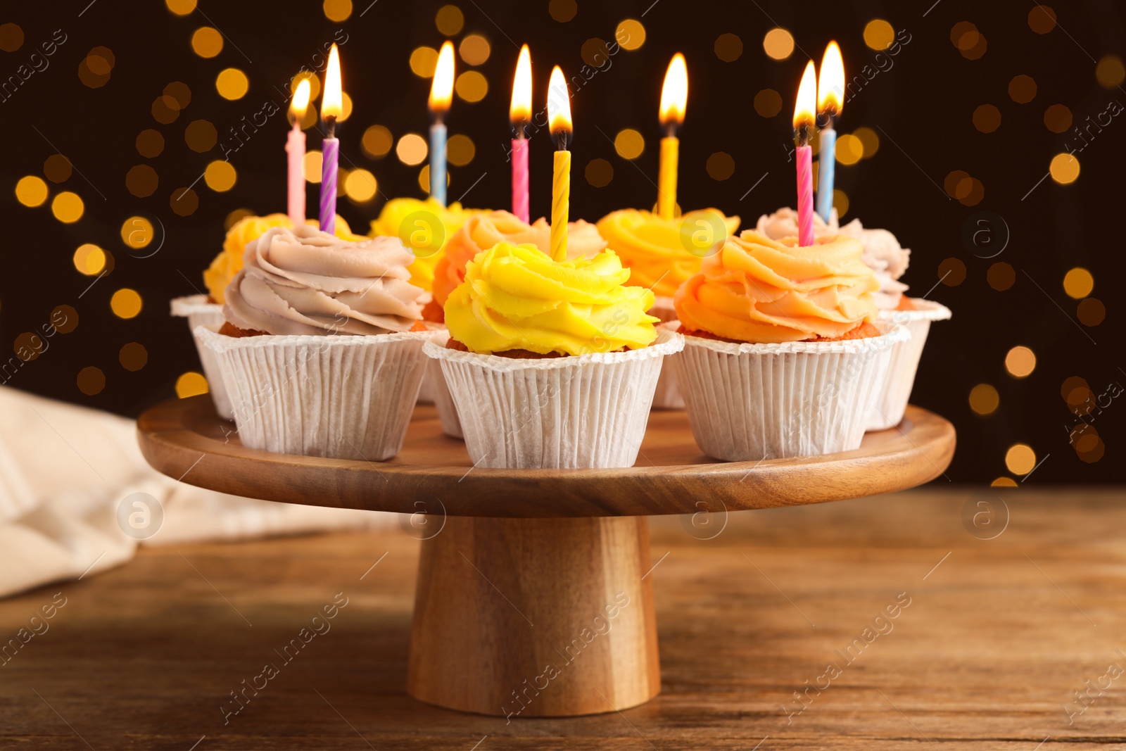 Photo of Stand with birthday cupcakes on wooden table against blurred lights