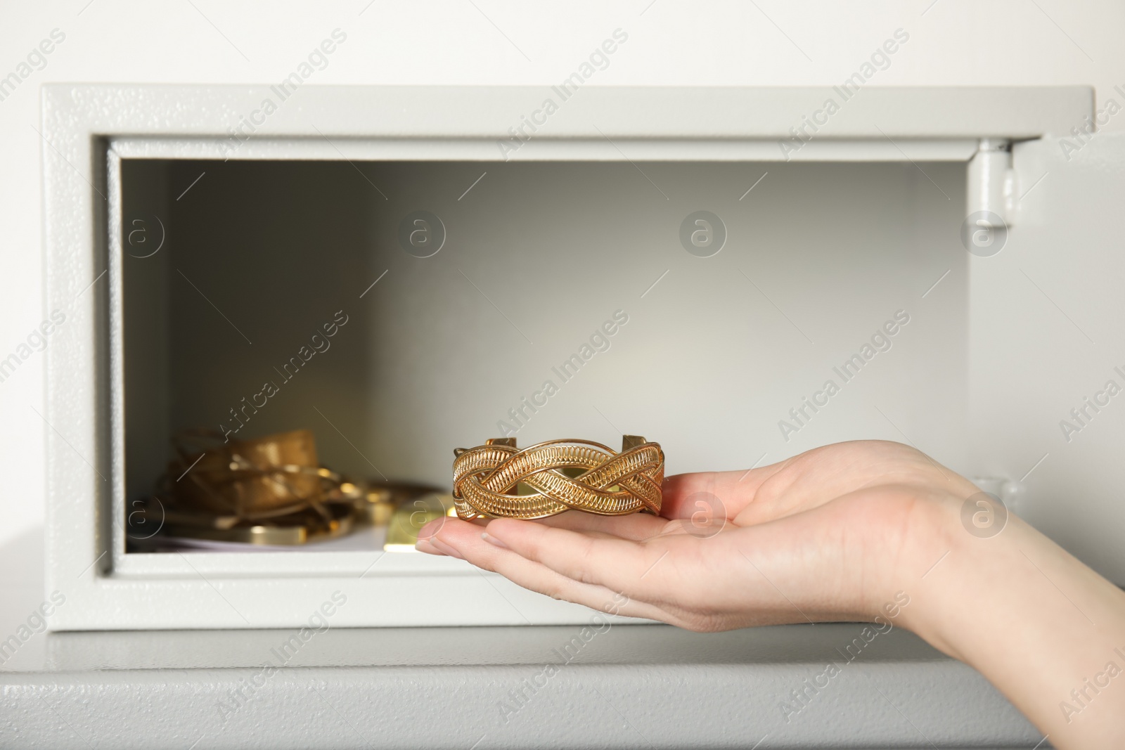 Photo of Woman taking gold bracelet out of steel safe on table against light background, closeup