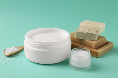 Photo of Moisturizing cream in open jars, sea salt and soap bars on turquoise background. Body care products