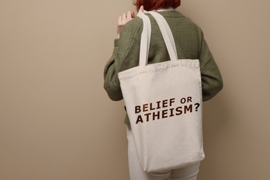 Woman holding bag with phrase Phrase Belief Or Atheism? on beige background, closeup