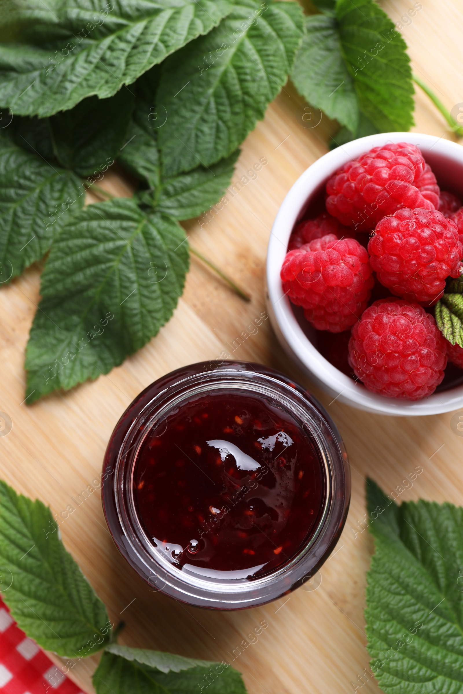 Photo of Jar of delicious raspberry jam, fresh berries and green leaves on wooden board, flat lay