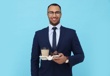 Photo of Happy young intern holding takeaway cup with hot drink and muffin on light blue background