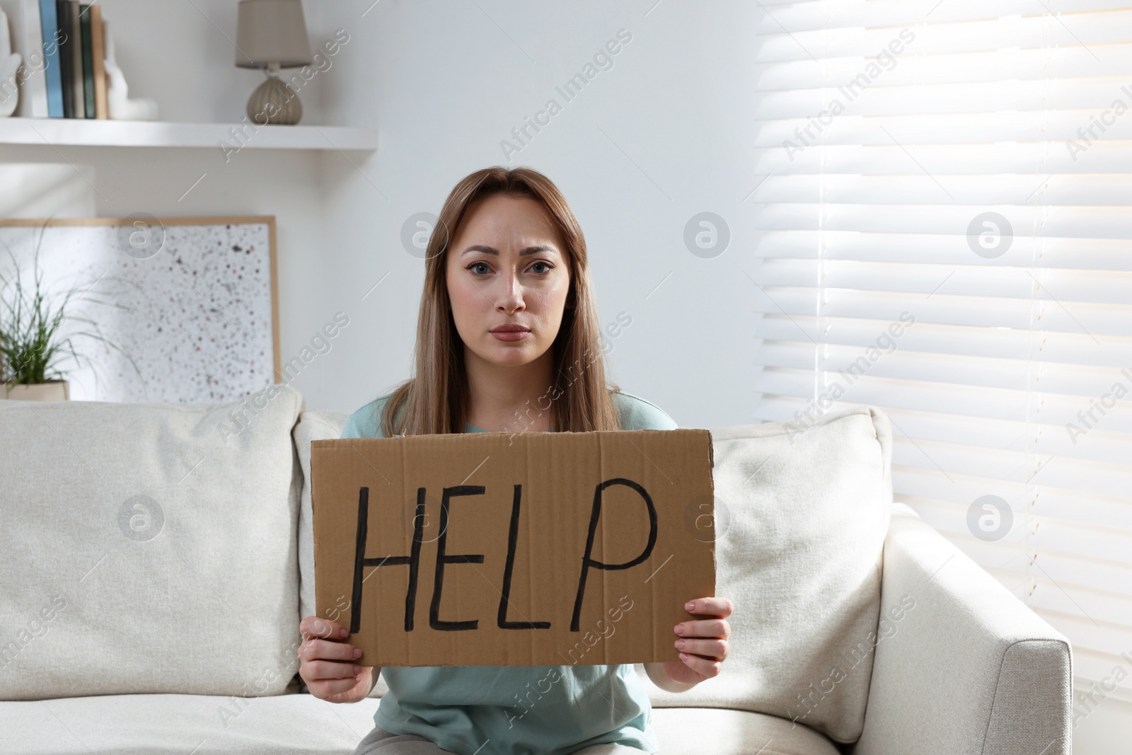 Photo of Unhappy young woman with HELP sign on sofa indoors