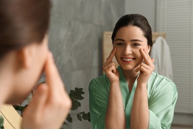 Photo of Young woman massaging her face near mirror in bathroom