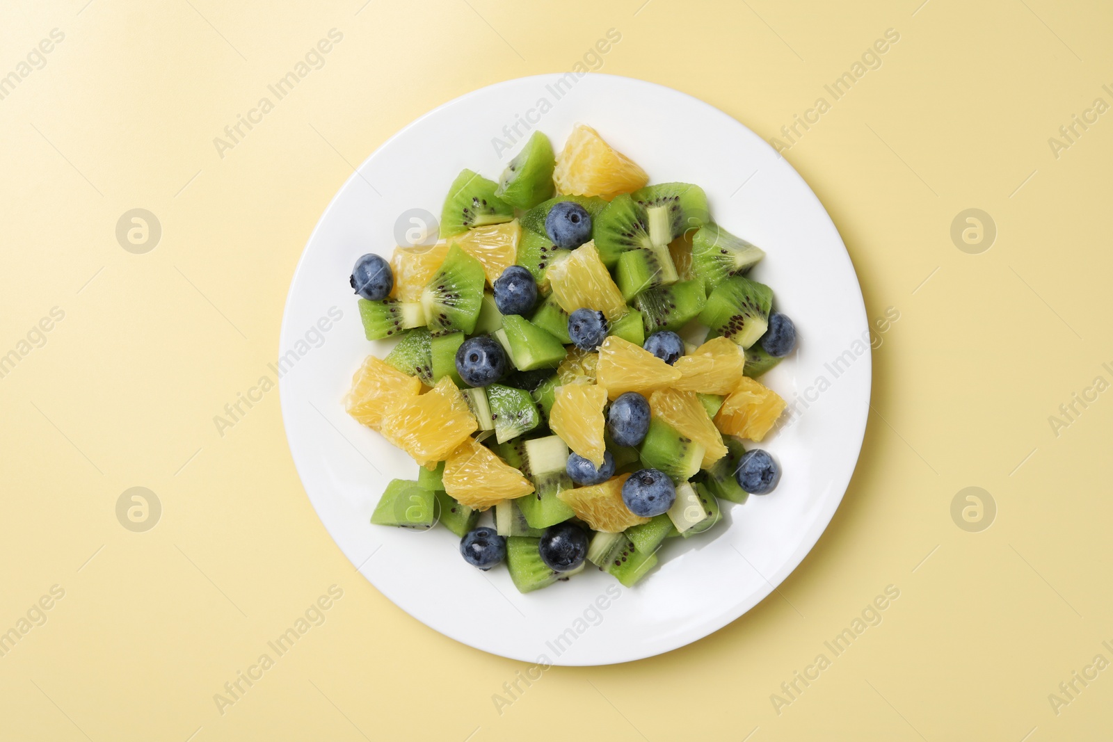 Photo of Plate of tasty fruit salad on pale yellow background, top view