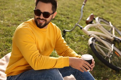 Young man sitting on green grass and holding takeaway coffee near bicycle outdoors