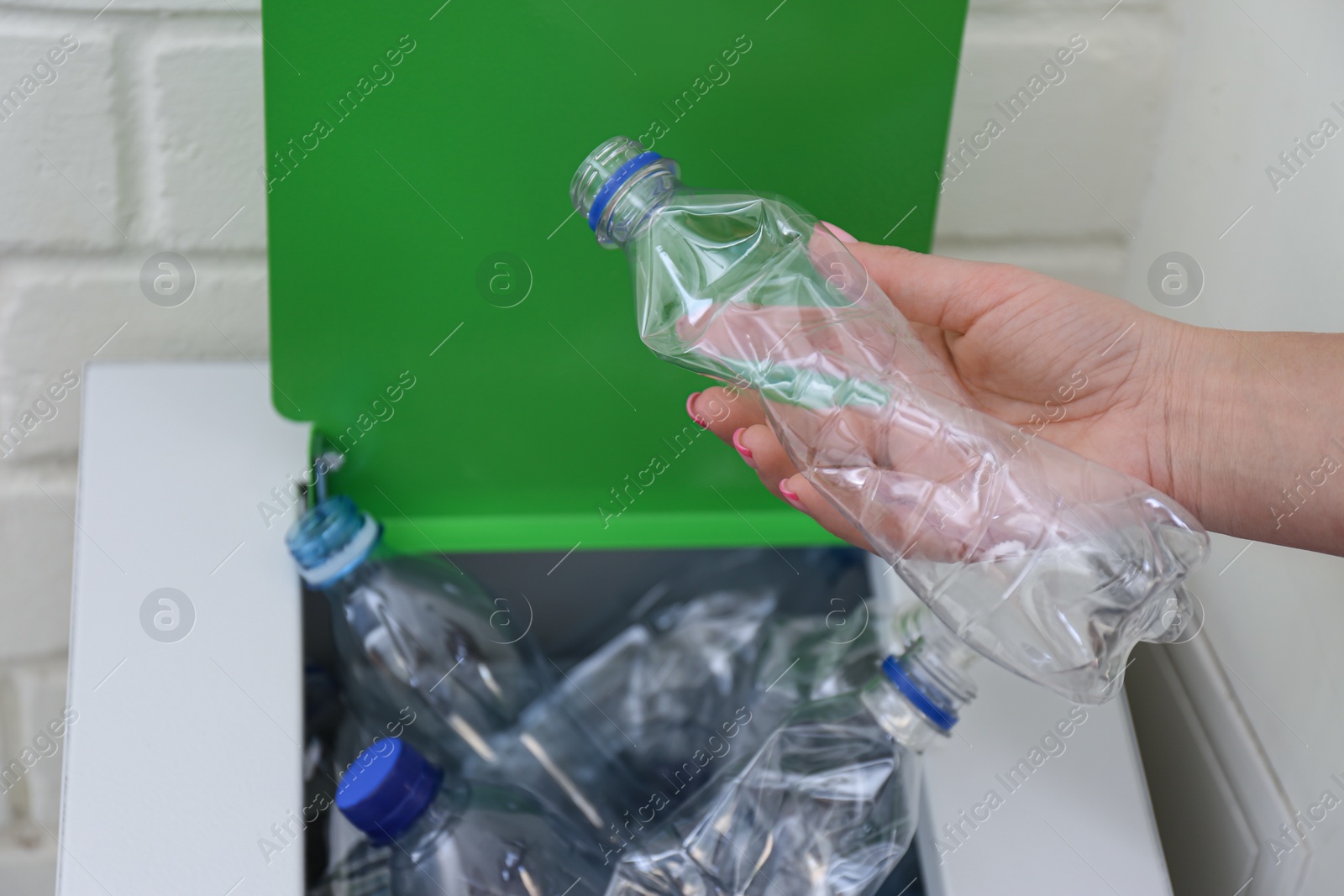 Photo of Woman putting used plastic bottle into trash bin near white brick wall, closeup. Recycling problem