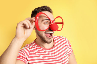 Photo of Emotional young man with party glasses and clown nose on yellow background. April fool's day