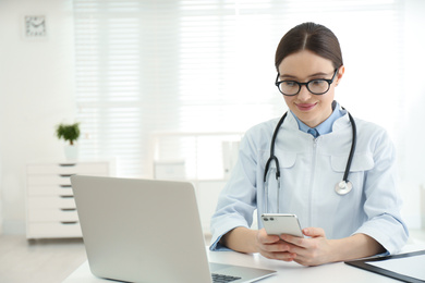 Young female doctor with smartphone at table in office