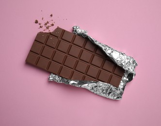 Photo of Bitten milk chocolate bar wrapped in foil on pink background, top view